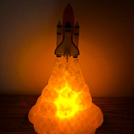 A glowing rocket ship lamp with a bright yellow and orange light emanating from a textured cloud base, set against a dark wall.