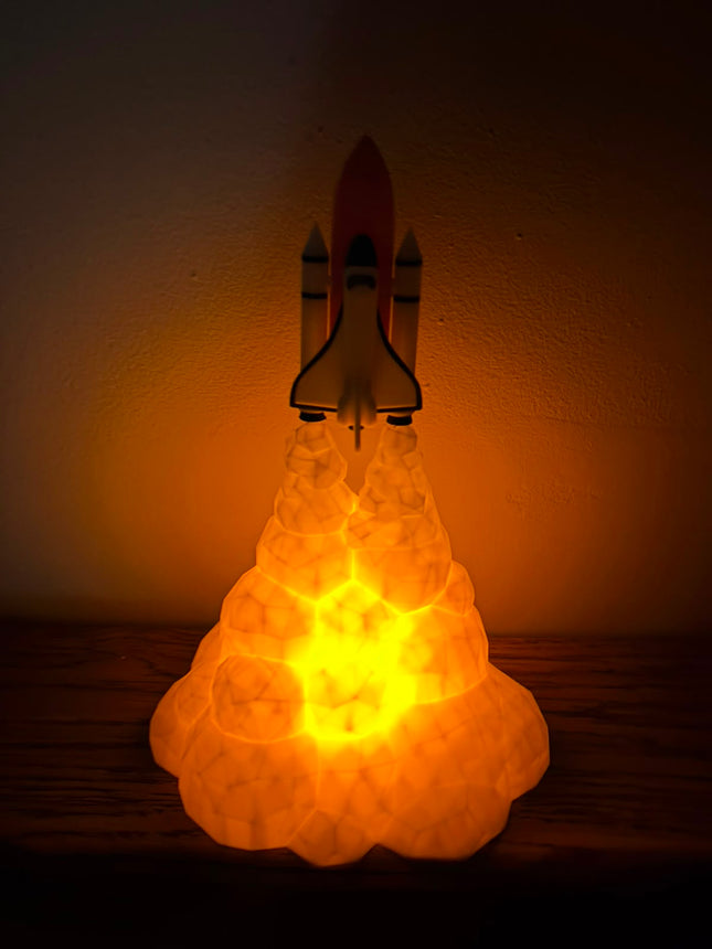 A glowing rocket ship lamp with a bright yellow and orange light emanating from a textured cloud base, set against a dark wall.